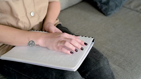 The-Camera-Focuses-On-Woman-Hand-Reading-A-Braille-Book-Sitting-On-The-Sofa-At-Home