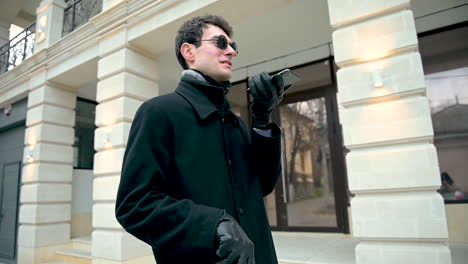 Unteransicht-Eines-Blinden-Mannes-Mit-Sonnenbrille-Und-Schwarzem-Mantel-Im-Freien,-Während-Er-Einen-Gehstock-Und-Ein-Smartphone-Hält-Und-Freihändig-Telefoniert