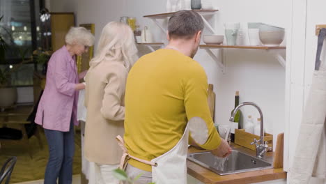 Rear-View-Of-A-Man-Washing-The-Family-Dinner-Dishes-At-The-Sink-In-The-Kitchen-While-Two-Mature-Women-Removing-The-Plates-From-The-Table