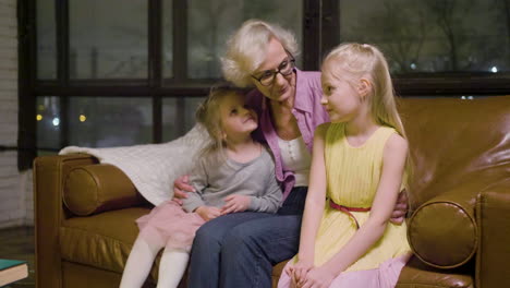 Grandmother-Hugging-To-Her-Two-Granddaughters-Sitting-On-The-Sofa-While-Talking-In-The-Living-Room-At-Home