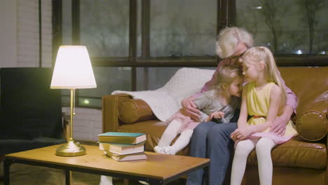 Grandmother-Hugging-To-Her-Two-Granddaughters-Sitting-On-The-Sofa-While-Talking-In-The-Living-Room-At-Home-1