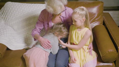 Top-View-Of-Grandmother-Hugging-To-Her-Two-Granddaughters-Sitting-On-The-Sofa-While-Talking-In-The-Living-Room-At-Home