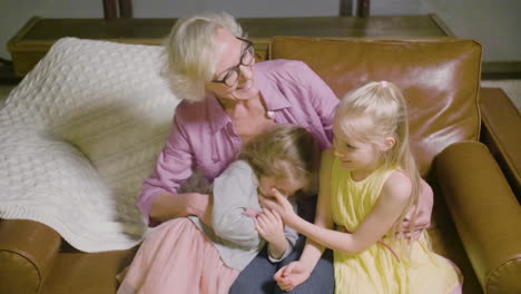 Top-View-Of-Grandmother-Hugging-To-Her-Two-Granddaughters-Sitting-On-The-Sofa-While-Talking-In-The-Living-Room-At-Home-1