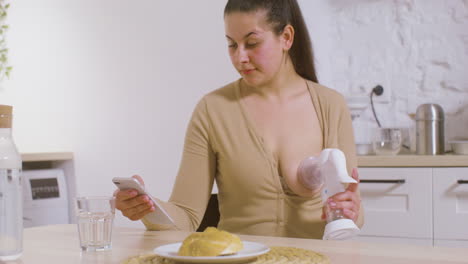 Young-Mother-Using-Breast-Pump-And-Scrolling-On-Mobile-Phone-While-Sitting-At-Table-At-Home