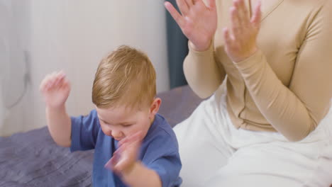 Happy-Young-Woman-Clapping-Hands-And-Playing-With-Her-Baby-Boy-While-Sitting-On-The-Bed-At-Home