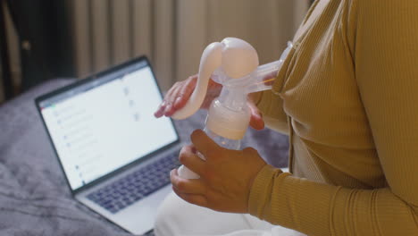 Close-Up-Of-A-Woman-Using-Breast-Pump-While-Sitting-On-The-Bed-With-Laptop-Computer-At-Home