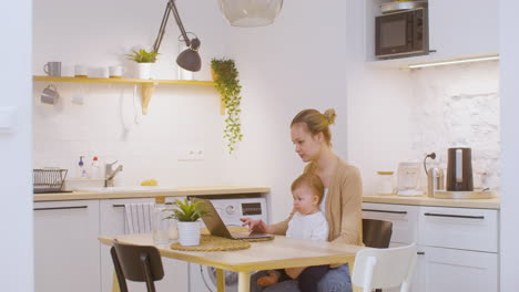 Mujer-Joven-Que-Trabaja-En-Una-Computadora-Portátil-Mientras-Está-Sentada-Con-Un-Bebé-En-Casa