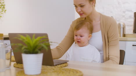 Young-Woman-Working-On-Laptop-Computer-While-Sitting-With-Baby-Boy-At-Home-1