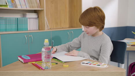 Concentrated-Ginger-Boy-Sitting-At-Desk-And-Writing-In-Notebook-During-English-Class-At-School