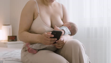 Joven-Madre-Amamantando-A-Su-Bebé-Recién-Nacido-Y-Usando-El-Teléfono-Móvil-Mientras-Se-Sienta-En-La-Cama-En-Casa