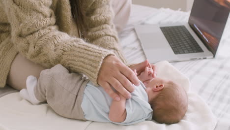 Unerkennbare-Mutter,-Die-Am-Laptop-Arbeitet-Und-Ihr-Neugeborenes-Baby-Kuschelt,-Während-Sie-Zu-Hause-Auf-Dem-Bett-Sitzt