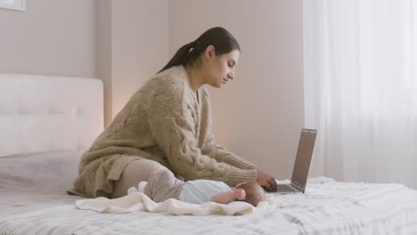 Junge-Mutter-Sitzt-Auf-Dem-Bett-Und-Arbeitet-Am-Laptop,-Während-Ihr-Neugeborenes-Baby-Neben-Ihr-Liegt