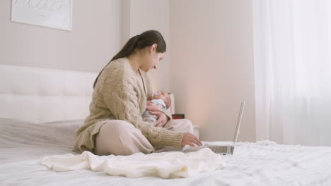 Brünette-Frau-Sitzt-Auf-Dem-Bett-Vor-Dem-Laptop,-Während-Sie-Ihr-Baby-Hält-Und-Ihn-Küsst