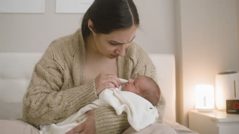 Front-View-Of-Brunette-Smiling-Woman-Sitting-On-The-Bed-Holding-Her-Baby-And-Caressing-Him