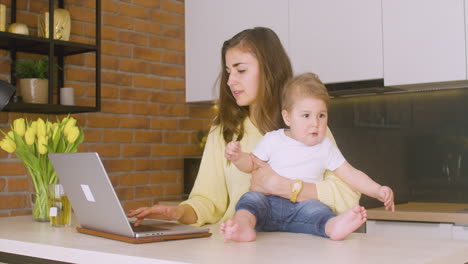 Mujer-Sosteniendo-A-Su-Bebé-Sentado-En-El-Mostrador-De-La-Cocina-Mientras-Usa-La-Computadora-Portátil
