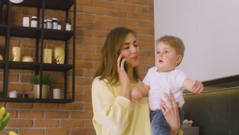 Woman-Holding-Her-Baby-In-The-Kitchen-While-Talking-On-The-Phone