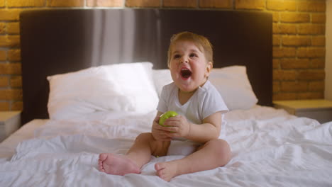 Front-View-Of-A-Baby-Sitting-On-The-Bed-In-Bedroom-While-Bitting-A-Green-Apple-1