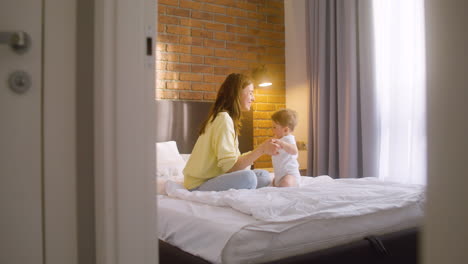 Woman-Helping-Her-Baby-Get-Up-And-Hugging-Him-While-Sitting-On-The-Bed-At-Home