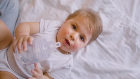 Draufsicht-Auf-Ein-Baby,-Das-Auf-Dem-Bett-Liegt-Und-Im-Schlafzimmer-In-Die-Kamera-Schaut,-Während-Es-Wasser-Aus-Der-Babyflasche-Trinkt