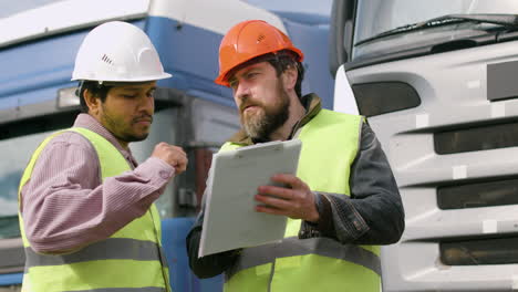 Worker-Wearing-Vest-And-Safety-Helmet-Organizing-A-Truck-Fleet-In-A-Logistics-Park-While-Consulting-A-Document-And-Holding-A-Smartphone-1