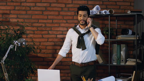 Good-Looking-Arabian-Businessman-Speaking-On-The-Phone-While-Standing-In-The-Office,-Then-Sits-At-The-Table-With-Laptop-Computer