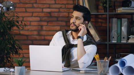 Junger-Arabischer-Geschäftsmann,-Der-Am-Laptop-Sitzt,-Am-Schreibtisch-Sitzt-Und-Fröhlich-Am-Telefon-Spricht