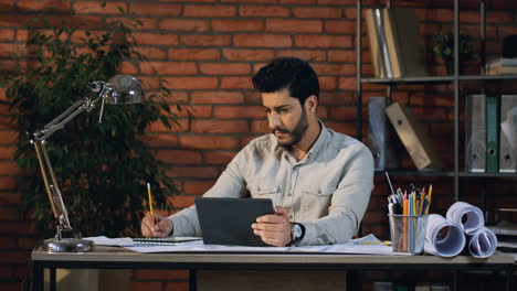Hombre-De-Negocios-árabe-Sentado-En-La-Mesa-De-La-Oficina,-Viendo-Algo-En-La-Pantalla-Del-Dispositivo-De-Tableta-Y-Escribiendo-Notas