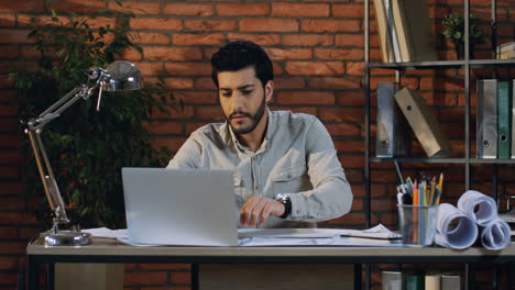 Young-Arabian-Architect-Drawing-Some-Plans-And-Looking-For-Example-On-The-Laptop-Screen-In-His-Modern-Office-Room