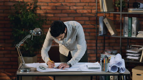 Young-Architect-Drawing-Some-Plan-Of-The-New-Building-On-The-Big-Sheet-Of-Paper-On-Desk-In-The-Urban-Cozy-Office