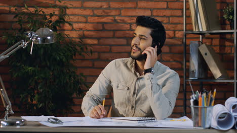 Young-Man-Architect-Talking-On-The-Phone-Cheerfully-While-Sitting-At-The-Desk-With-Drawing-On-It