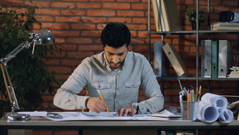 Arabian-Architect-Drawing-Some-Building-Plan-On-Desk-In-His-Office,-Then-Rises-Up-His-Head-And-Smiling-To-The-Camera