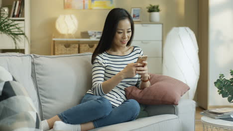 Charming-Young-Woman-Wearing-Striped-Blouse,-Tapping-And-Texting-On-The-Smartphone-Sitting-On-A-Sofa-In-The-Living-Room