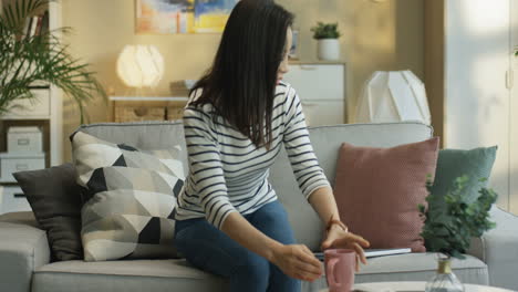 Young-Woman-Wearing-A-Striped-Blouse-Walking-Over-To-The-Sofa-And-Sitting-Down