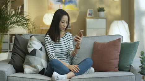 Young-Woman-In-Striped-Shirt-And-Headphones-Listening-To-Music-On-Smartphone-Sitting-On-Sofa-In-The-Cozy-Living-Room