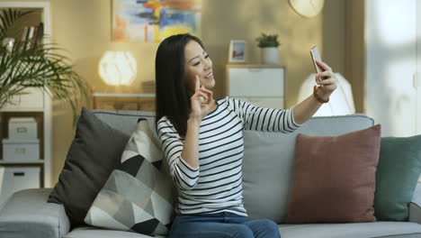 Young-Woman-In-Striped-Shirt-Posing-To-The-Smartphone-Camera-And-Smiling-While-Taking-A-Selfie-Photos-Sitting-On-Couch-In-The-Living-Room
