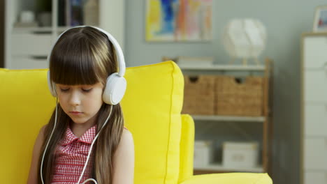 Little-Pretty-Girl-Wearing-Big-Headphones-Sitting-On-Yellow-Sofa-In-Living-Room-At-Home
