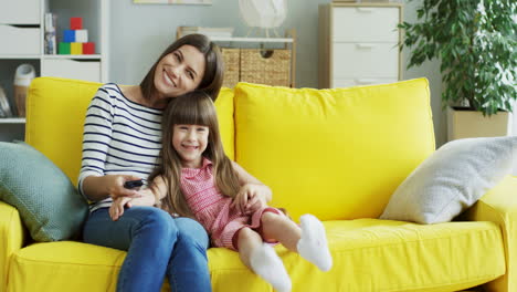 Young-Beautiful-Mother-Hugging-Her-Small-Daughter-While-They-Sitting-On-Couch-In-The-Living-Room,-Turning-On-Tv-With-Remote-Control-And-Girl-Being-Very-Cheerful-And-Happy