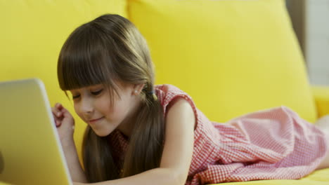 Little-Cute-Girl-Playing-With-Laptop-While-She-Is-Lying-On-Yellow-Couch-In-Living-Room