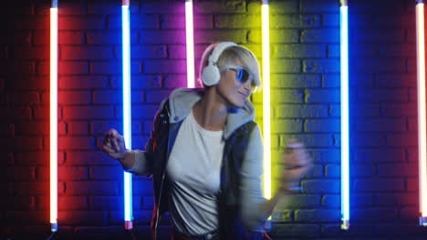 Blonde-Frau-Mit-Sonnenbrille-Und-Weißen-Kopfhörern-Tanzt-In-Einem-Raum-Mit-Bunten-Neonlampen-An-Der-Wand