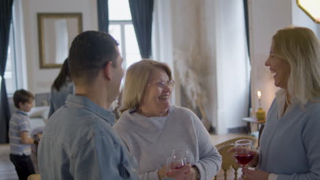 Two-Happy-Middle-Aged-Women-And-Man-Standing-And-Talking-At-Family-Party-While-Holding-Glasses-And-Having-Fun