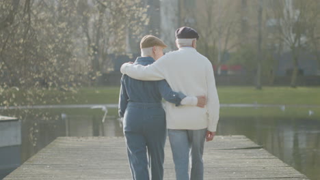 Back-View-Of-Elderly-Couple-Walking-In-Step-Along-Wooden-Pier-In-City-Park