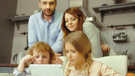 Boy-And-Girl-Siting-In-Kitchen-Using-Laptop-Showing-Something-To-Their-Parents