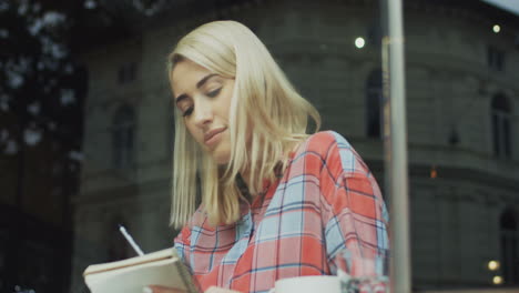 Blonde-Woman-Sitting-In-A-Cafe-And-Thinking-While-Writing-Something-With-A-Pencil-In-Her-Notebook