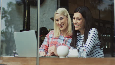 Two-Pretty-Women-Sitting-In-Front-Of-A-Laptop-Computer-And-Looking-At-Something-Interesting-In-The-Cafe-And-Having-A-Video-Call