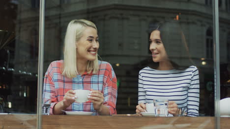 Portrait-Of-Two-Female-Friends-Drinking-Coffee-And-Smiling-To-The-Camera-While-Sitting-In-A-Cafe