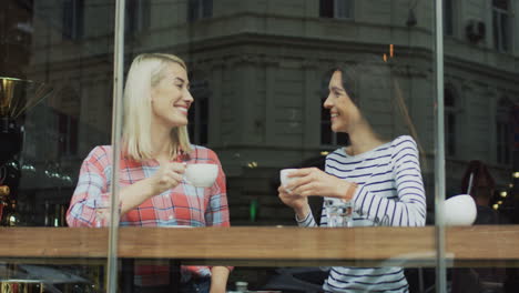Two-Smiling-Friends-Sitting-In-A-Cafe-Behind-A-Window,-Taking-And-Drinking-Coffee