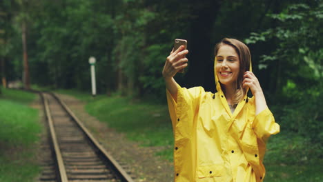 Süße-Frau-In-Einem-Gelben-Regenmantel,-Die-Selfie-Fotos-Auf-Einem-Smartphone-Macht-Und-Posiert