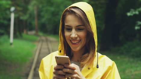 Close-Up-Of-Cute-Woman-In-A-Yellow-Raincoat-Standing-In-The-Middle-Of-The-Woods,-Laughing-And-Texting-On-Her-Smartphone