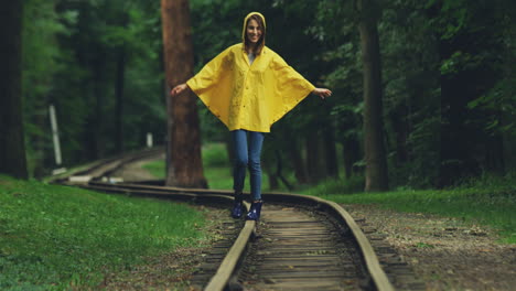 Chica-Atractiva-Con-Un-Impermeable-Amarillo-Caminando-Con-Cuidado-Por-El-Antiguo-Ferrocarril-En-El-Bosque
