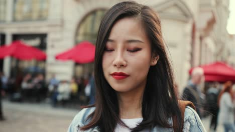 Pretty-Young-Woman-With-Red-Lips-Looking-Straight-To-The-Camera-And-Smiling-At-The-City-Square-With-Cafes-Umbrellas
